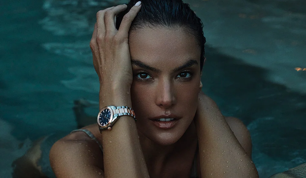 Model Alessandra Ambrosio poses in a body of water, showcasing the new 38 mm Seamaster Aqua Terra 150M. She has wet hair and rests her hands on her head, highlighting the watch on her wrist.