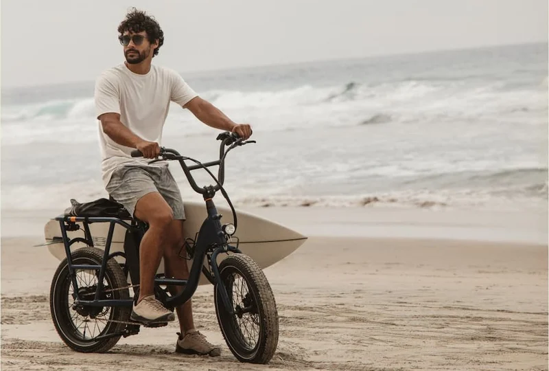 Man riding a bike on the beach with a surfboard, wearing casual coastal outfit under headline “The all-day explorer.”