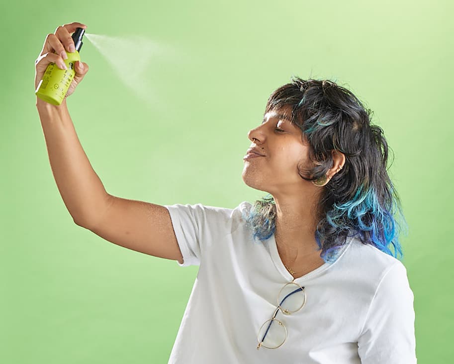 A person with colorful blue hair and a white shirt sprays toner on their face