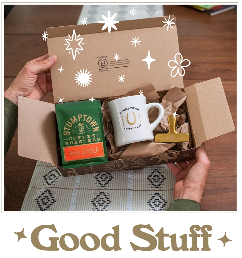 Open subscription box revealing Stumptown Coffee Club items: a green Hair Bender whole bean coffee bag, a white mug with horseshoe logo, and a gold bag clip nestled in brown packing paper. White star doodles decorate the image. Text below reads: “Good Stuff.”