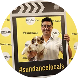 Man holding a dog in a picture frame. On the picture frame, there is #sundancelocals on it.
