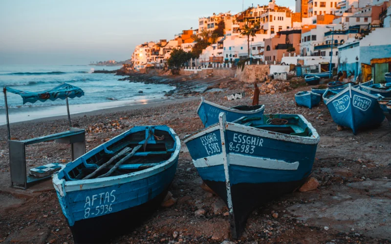 Two boats sit on a beach with Taghazout, Morocco, in the background.