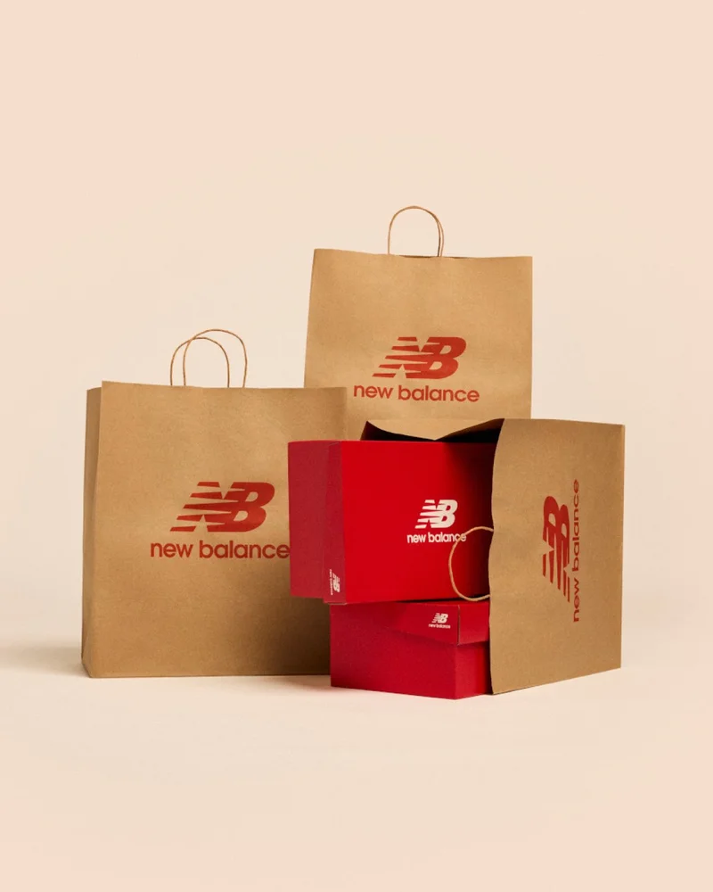 A stack of New Balance shoe boxes in a shopping bag against a pale apricot backdrop