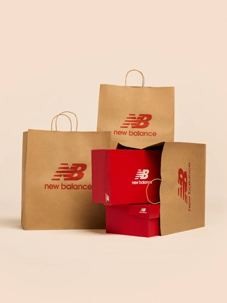 A stack of New Balance shoe boxes in a shopping bag against a pale apricot backdrop
