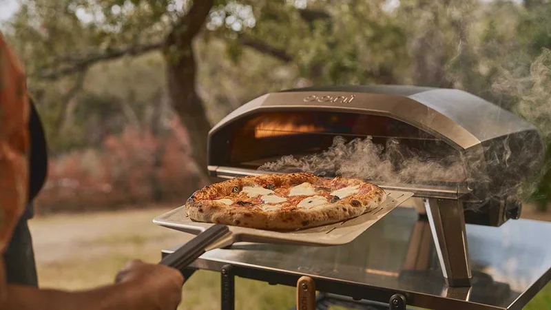 Ooni Koda 2 Pro on an Ooni table with a cooked pizza being taken out with an Ooni pizza peel