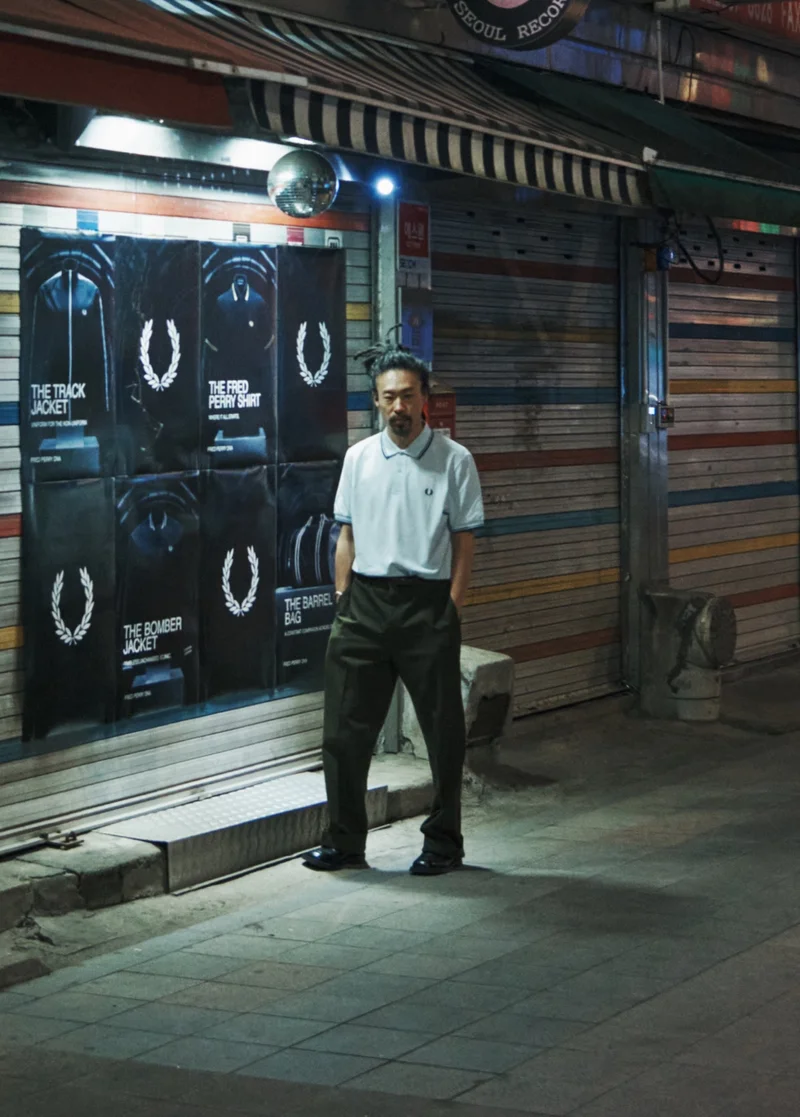 A Korean man stands in the distance on a dimly lit street in Soeul in front of Fred Perry posters and wears the Fred Perry Shirt in White / Light Ice / Navy. His long hair is tied back and his hands are tucked into the pockets of his black tailored trousers.