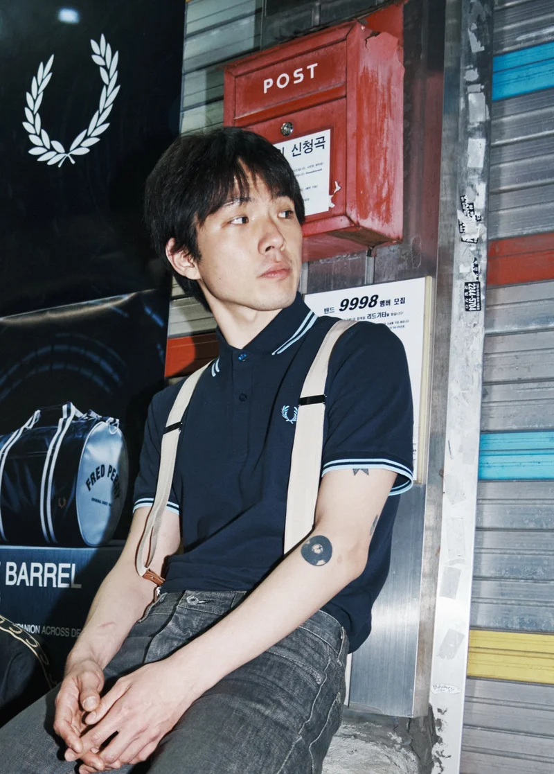 A Korean man with short straight hair sits against a wall on a street in Soeul. His hands sit comfortably in his lap, and he wears a Fred Perry Shirt in Navy / Light Ice / Light Ice. Braces attached to his grey jeans sit over his shoulders as he looks wistfully away from the camera into the distance.