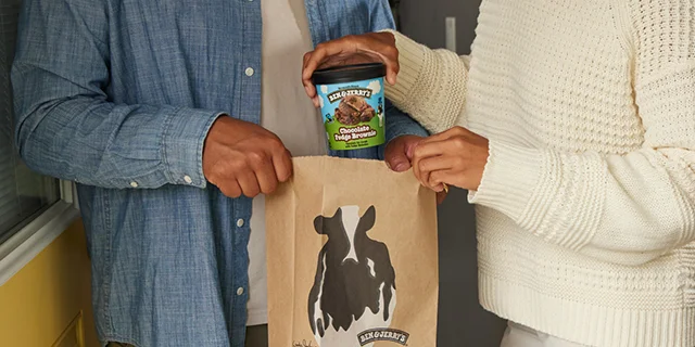 Two people holding a brown paper bag with the Ben & Jerry's cow on it. One person is lifting a pint of Ben & Jerry's Chocolate Fudge Brownie ice cream out of the bag.