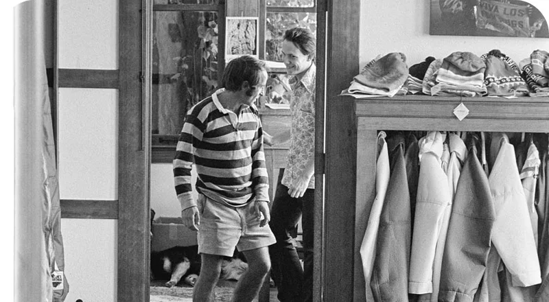 A black and white photo of two people walking through the door of an office next to a rack of hanging jackets.