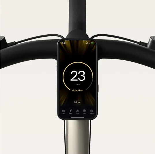 A render showing a smartphone mounted on the handlebars of a Cowboy e-bike, displaying the speed with an “Adaptive” mode indicator on the screen.