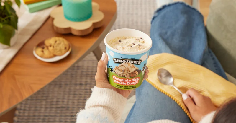 Hand holding a pint of Ben & Jerry's Chocolate Chip Cookie Dough ice cream in a home setting.