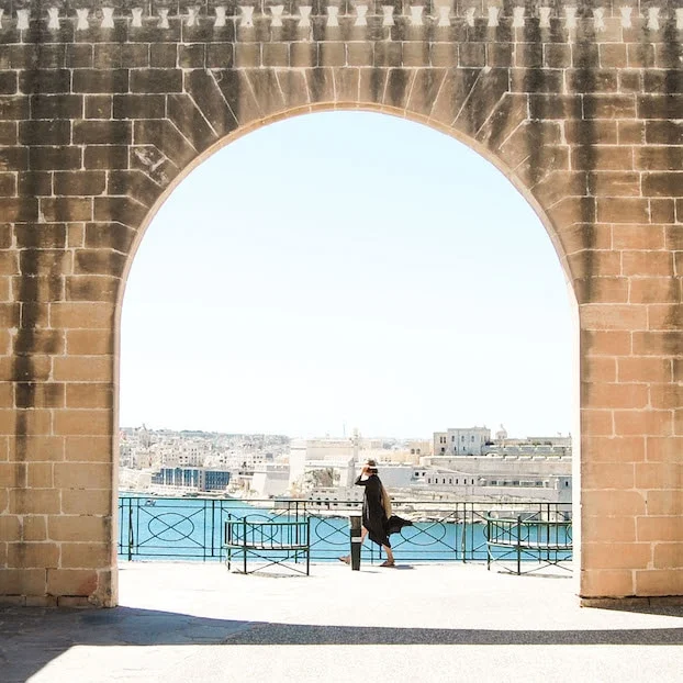A view through a brick wall's arched opening. A person in a black dress walks past a body of water and white buildings.