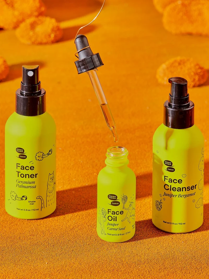 Three green bottles of skincare products stand on a sandy ground. A dropper full of oil is suspended by a wire.