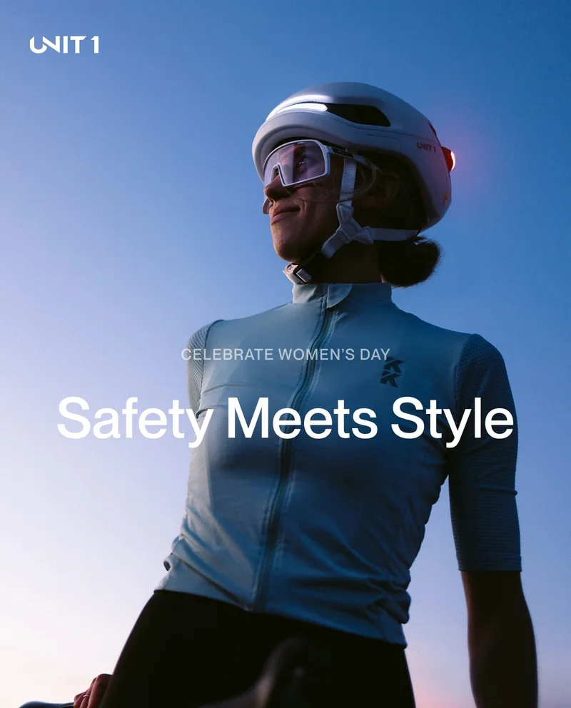 A woman wearing the UNIT 1 AURA smart helmet, illuminated by built-in front and rear lights, standing confidently against a twilight sky. Text overlay: "Celebrate Women's Day – Safety Meets Style."