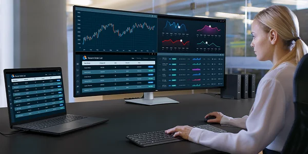 An office worker tracking the status of recent orders across multiple dashboards on a widescreen Dell UltraSharp 52 TBT monitor, laptop, wireless keyboard and mouse.