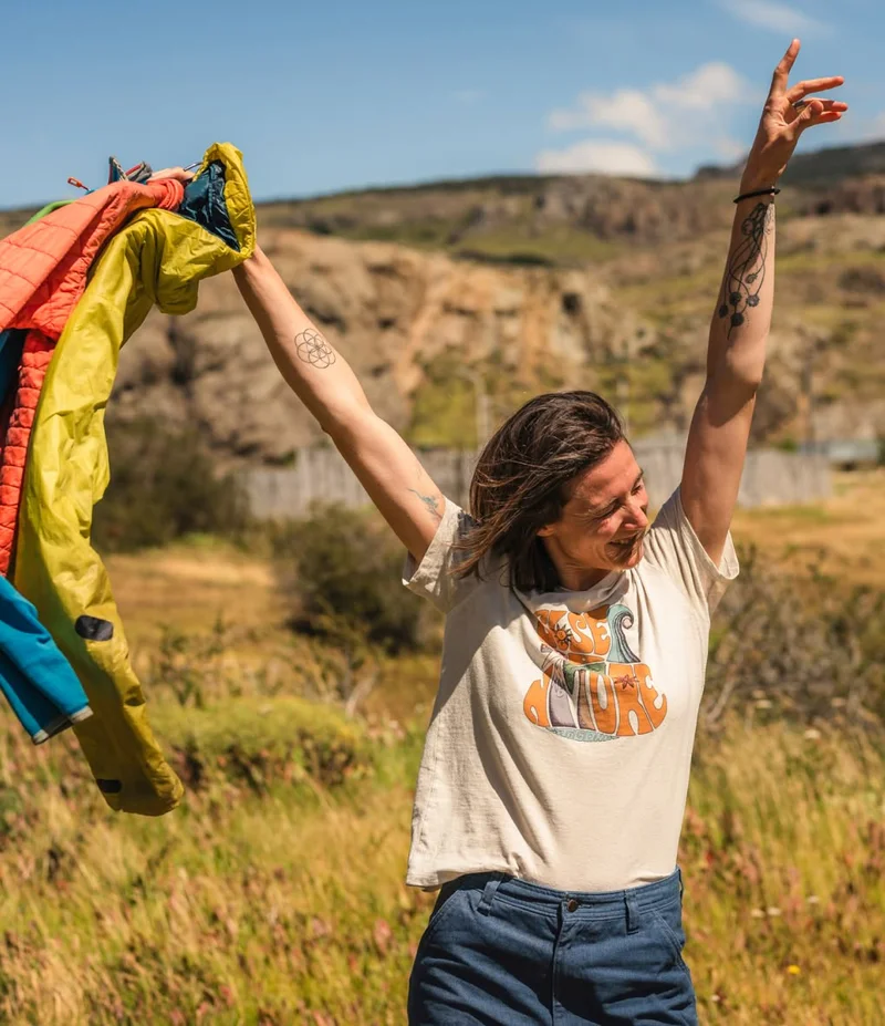 Let it go. A person stands in a meadow with arms raised and holds a few jackets in one hand.