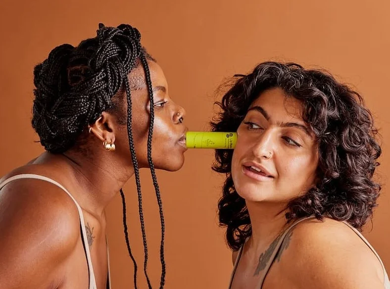 Two people balance a green tube of lip balm between their faces, one has the tube pressed against their lips while the other has the tube pressed against their cheek.