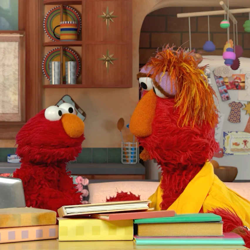 Elmo and a larger red, shaggy-haired muppet in a yellow robe face each other across a table piled with books in a colorful kitchen setting.