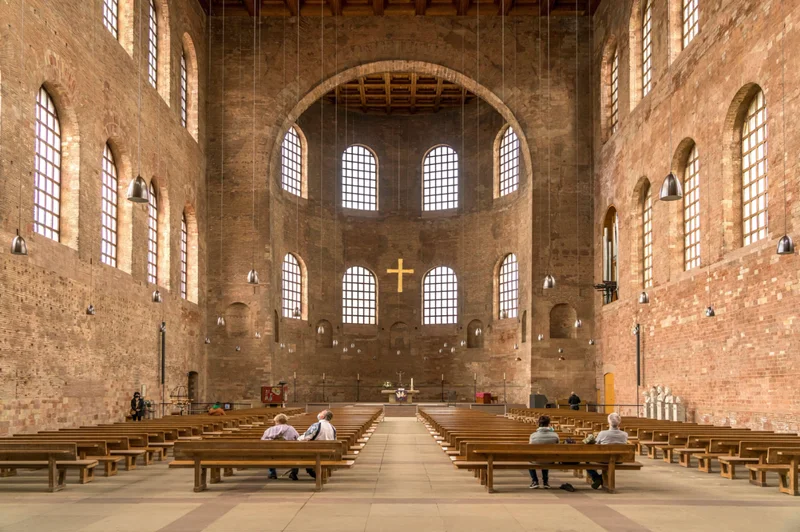 View of the inside of a large cathedral from the entrance. There are pews on the left and right with several people sitting in them. There is an altar in the back and a gold cross hanging halfway from the ceiling.