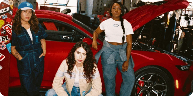 Three women wearing Ford x Sydney Sweeney workwear, in front of a red 2024 Ford Mustang® coupe