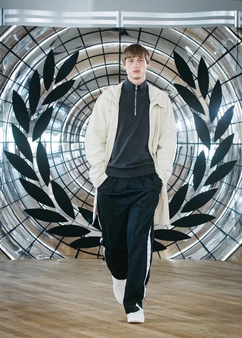 White male model with bowl cut hair walks confidently towards the camera on our runway, wearing a full black outfit with a slouchy beige parka jacket