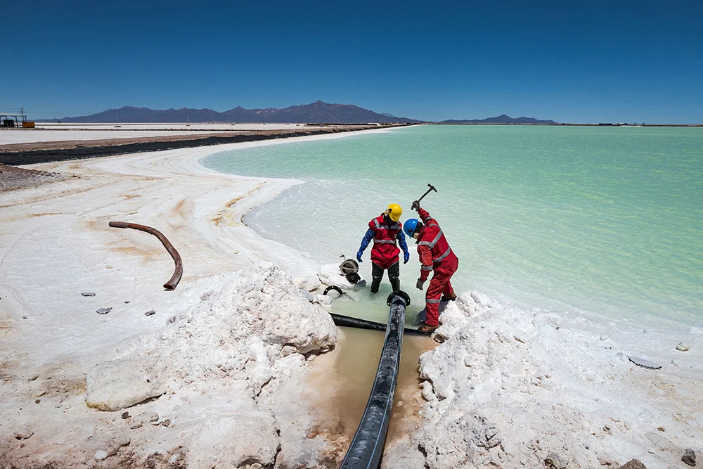 Workers, wearing red, one piece suits, at the lithium pilot plant use hammers to break up a layer of salt that periodically clogs the pipes transporting brine with lithium into large, turquoise blue evaporation pools.