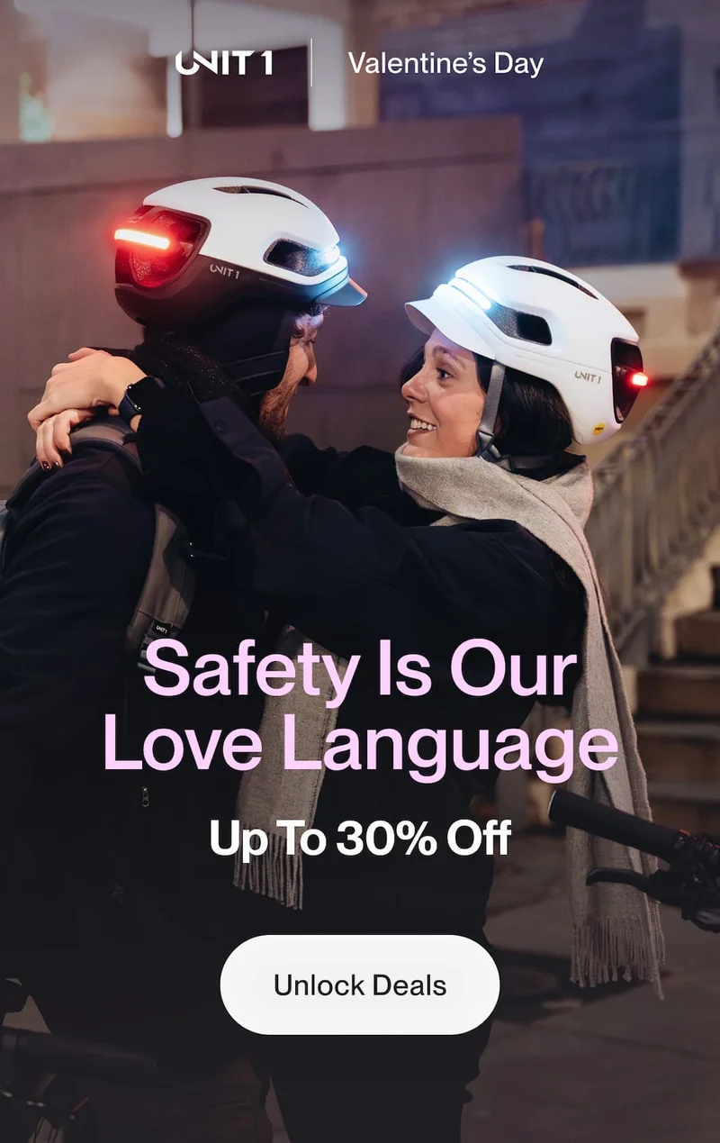 Couple wearing UNIT 1 smart helmets with front and rear lights illuminated, embracing beside their bikes in an urban setting at night.