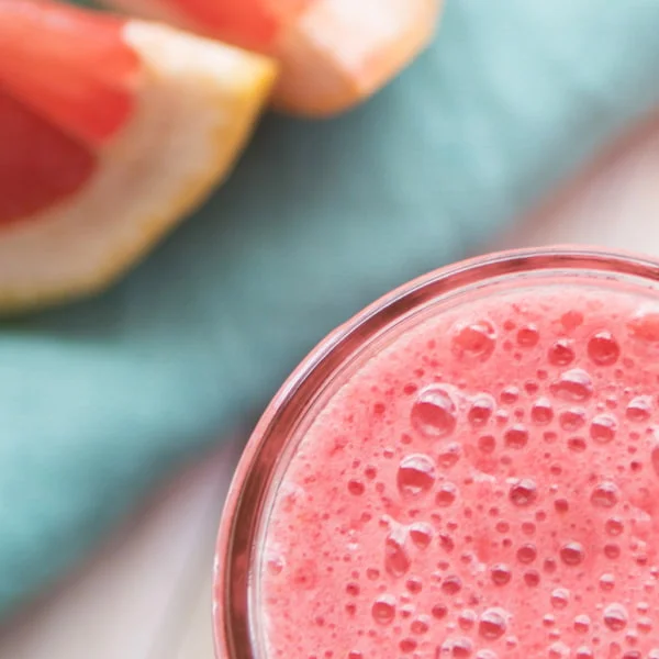 A glass of pink smoothie with grapefruit slices in the background on a turquoise cloth.