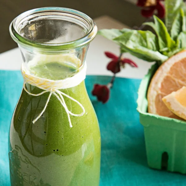 A glass bottle of green smoothie tied with a string, next to a carton of grapefruit slices and basil leaves on a turquoise surface.
