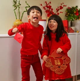Two children wearing red outfits smile joyfully, holding festive decorations. The background features a cabinet with vases and red ornaments, suggesting a celebration.