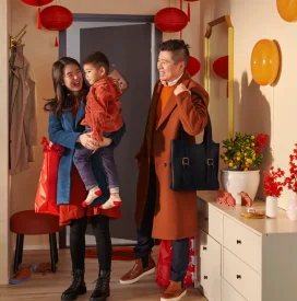 A joyful family enters a festive room, adorned with red lanterns and decorations. They are warmly dressed, holding a child, conveying happiness and celebration.