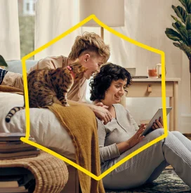 A couple smiling, sitting in a cozy room using a tablet. One person lies on a bed with a cat nearby. Warm lighting and a relaxed, happy atmosphere.