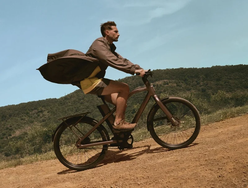rider on a Cowboy Cross e-bike glides over a rugged dirt path, their coat billowing behind them. The bike’s front suspension absorbs the terrain, highlighting its off-road capabilities.