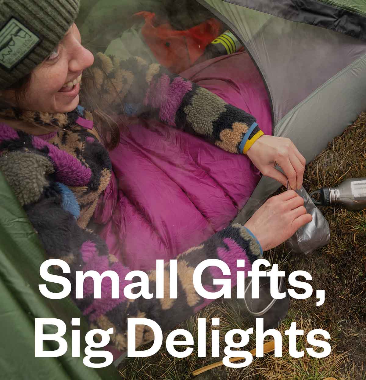 A person laughing in their sleeping bag while fixing a cup of coffee just outside the tent door.