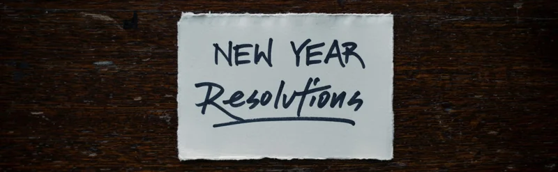 A white card that says “New Year Resolutions” in black ink on a dark wood desk with a pen underneath.