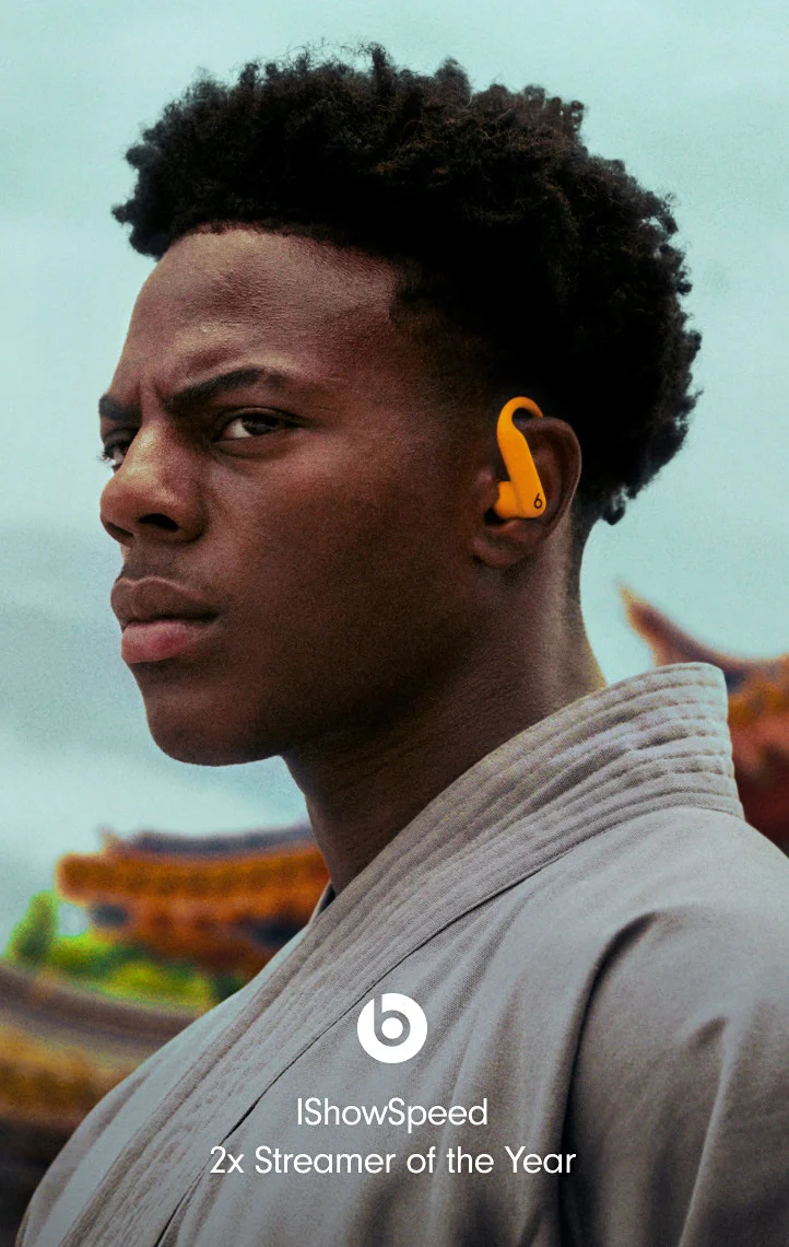 Online streamer IShowSpeed, a young man, wears orange Powerbeats Pro 2 earbuds and a light-colored robe, looking seriously to the left.
