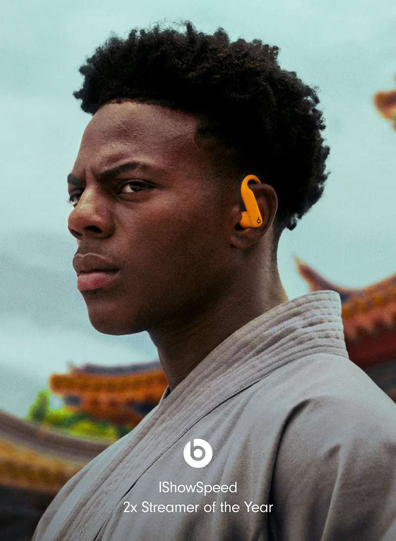 Online streamer IShowSpeed, a young man, wears orange Powerbeats Pro 2 earbuds and a light-colored robe, looking seriously to the left.