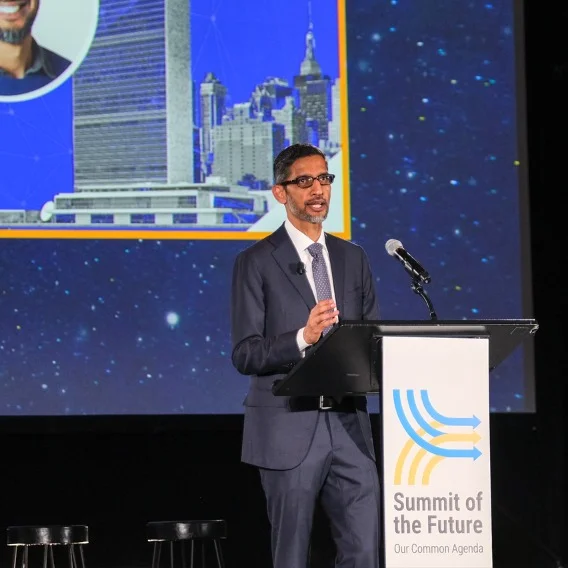 A photo of Sundar Pichai giving a presentation on a stage, standing behind a podium with a banner that reads 'Summit of the Future.”