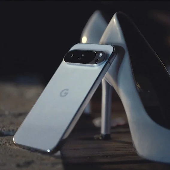 A white Google Pixel phone stands upright, leaning against a pair of white high heels on a dark wooden surface.