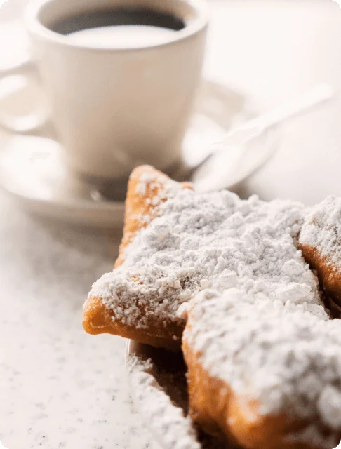 Image Description: Two plates of beignets covered in powdered sugar with a cup of coffee on a countertop