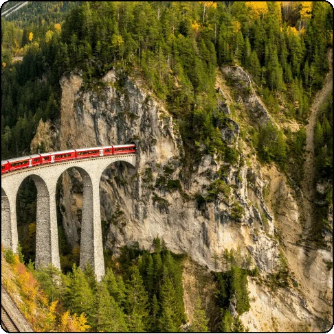 Image Description: A red train cuts through a woodsy gorge and disappears into the side of a mountain.