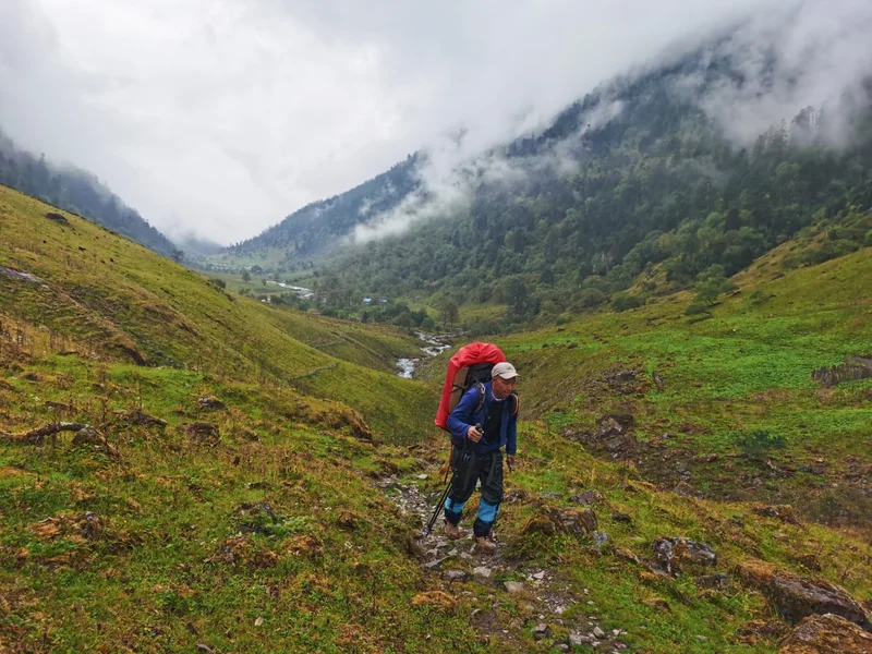A man wearing a bright red backpack hikes across a lush green trail surrounded by green mountains. Behind him are clouds that cover the top of the mountain peaks.