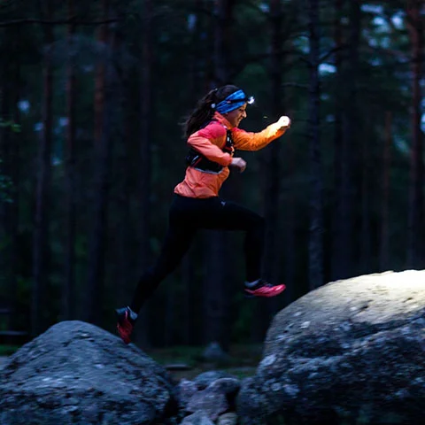 A woman jumps over large rocks at nighttime while wearing a headlamp, lighting up the rocks below.