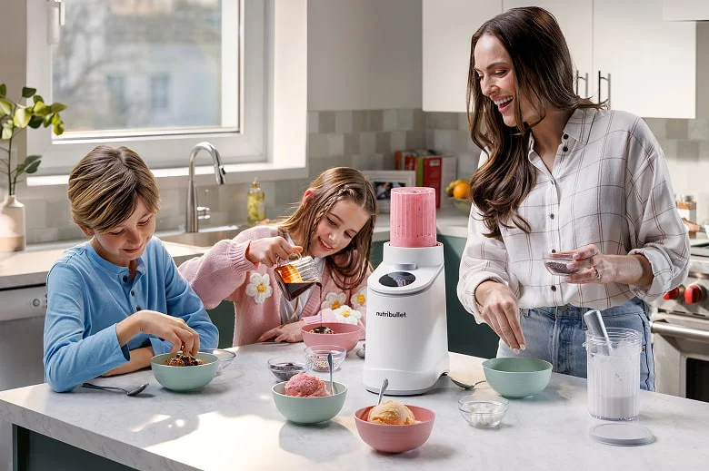 A woman and two children are gathered around a white kitchen island eating ice cream with a white nutribullet chill in the center filled with a pink frozen mixture.