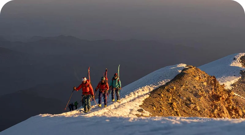 Three backcountry skiers walk along a snowy ridge with their skis on their backs.