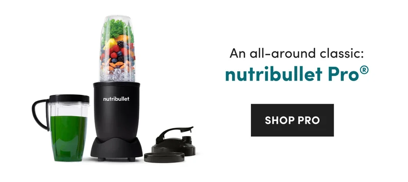 A black nutribullet personal blender filled with kale, fruit, almonds, and ice next to a cup of green liquid on a white background.