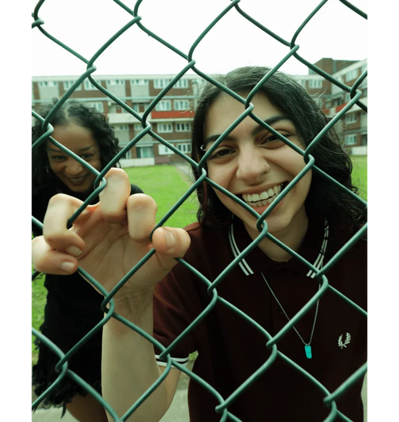 Two young women laughing behind a wired community fence wearing Fred Perry Shirts
