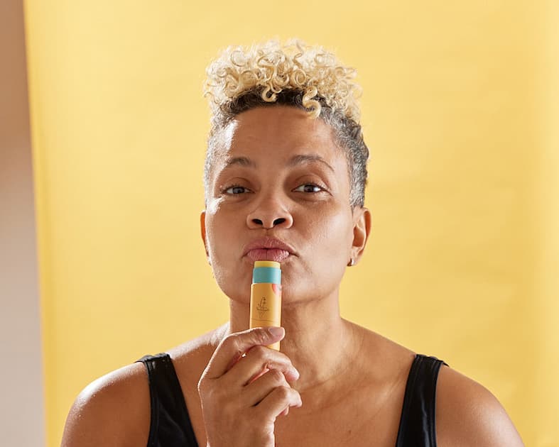 A light-skinned Black person with blond short curly hair puckering up over an orange lip balm tube