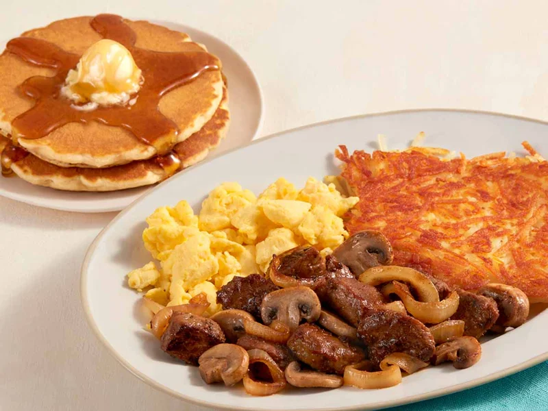 A plate of scrambled eggs, hash browns, steak tips and mushrooms, with a plate of two buttermilk pancakes with syrup and butter.