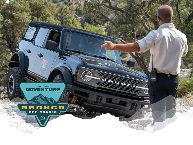 An instructor guiding a man who is driving a Ford Bronco® SUV on rocky terrain overlaid with the Bronco Off‑Roadeo® Half‑Day Adventure logo in the bottom left corner